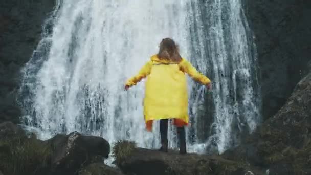 Young woman arms outstretched behind spectacular waterfall in Iceland. Shot of young woman outstretching arms by majestic waterfall. Success and achievement concept — Stock Video