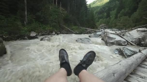 Ένας άντρας κάθεται σε μια ξύλινη γέφυρα, και τα πόδια κρέμονται κάτω. Ένα γρήγορο ορεινό ποτάμι ρέει. Ταξίδια στο βουνό. — Αρχείο Βίντεο