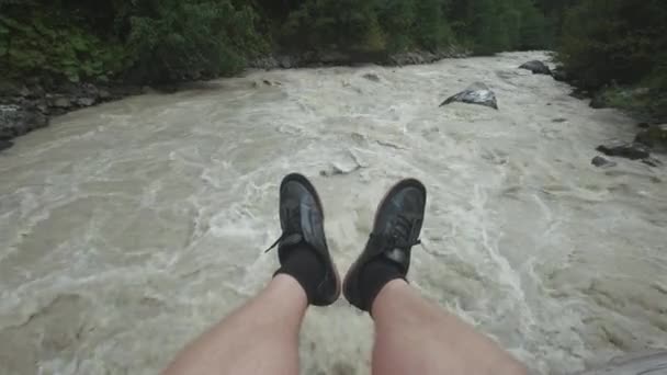 一个男人坐在一座木桥上，双腿悬着下来。一条湍急的山河流淌着。山地旅行. — 图库视频影像