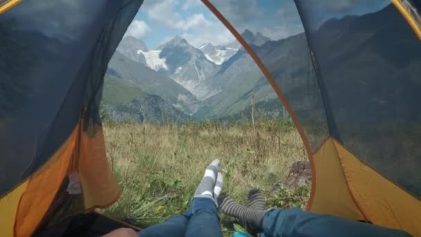 Vista de dentro de uma tenda nas montanhas. Um par de homens e mulheres viajantes jazem numa tenda. Faça uma pausa da trilha de caminhada ao longo da rota da montanha. Os pés saem da tenda . — Vídeo de Stock