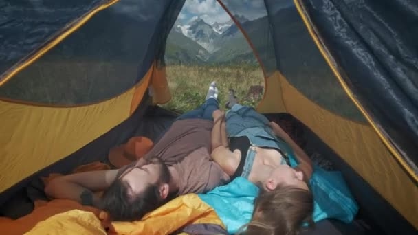 Uitzicht vanuit een tent in de bergen. Een paar man en vrouw reizigers liggen in een tent. Neem een pauze van het wandelpad langs de bergroute. Voeten steken uit de tent. — Stockvideo