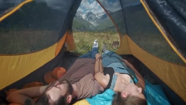 Widok z wnętrza namiotu w górach. Kilka osób podróżujących mężczyzna i kobieta leżą w namiocie. Pospacerować po szlaku górskim wzdłuż szlaku górskiego. Stopy trzymać się z namiotu. — Wideo stockowe