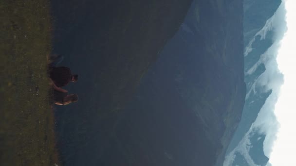 Vertikální záběr: mladý pár sedí na úbočí kopce a dívá se na západ slunce a hory. Koncept cestování a jednoty s přírodou. Zasněžené hory. — Stock video