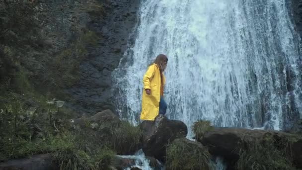 Młoda dziewczyna wędrowiec w żółtym płaszczu idzie do wodospadu w górach, podnosi ręce do góry, cieszy się przyrodą i życiem. Podróżowanie w górach, przygoda w podróży. Koncepcja stylu życia — Wideo stockowe
