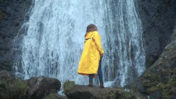 Jonge meisjeswandelaar in een gele regenjas loopt naar een waterval in de hooglanden, steekt zijn handen op, geniet van de natuur en het leven. Reizen in de bergen, avontuur in reis. Levensstijl concept — Stockvideo