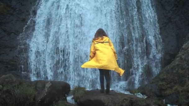 Молодая девушка путешественник в желтом плаще прогулки к водопаду в горах, поднимает руки вверх, наслаждается природой и жизнью. Путешествие в горы, приключение в поездке. Концепция образа жизни — стоковое видео