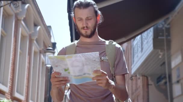 Ένας νεαρός με μούσι, ένας τουρίστας ταξιδιώτης περπατάει σε μια ευρωπαϊκή πόλη με ένα χάρτη στα χέρια του. Ψάχνω για έναν τρόπο για αναψυχή. Κοιτάζοντας την πόλη στο δρόμο της πόλης. Ευρώ-ταξίδι. — Αρχείο Βίντεο