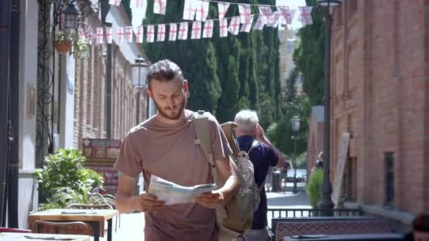 Um jovem com barba, um viajante turístico está andando em uma cidade europeia com um mapa em suas mãos. À procura de uma forma de recreação. olhando ao redor na rua da cidade. Euro-viagem . — Vídeo de Stock