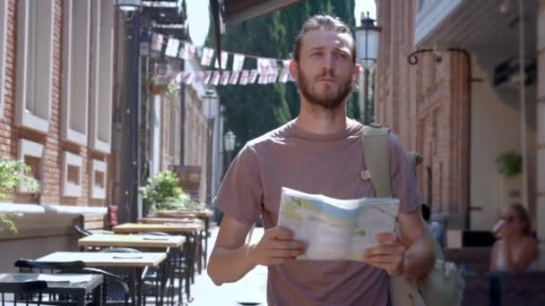 Egy fiatal fickó egy szakállas, egy turista utazó sétál az európai város egy térképet a kezében. Keresi a módját, hogy kikapcsolódás. keresett körül a város utcán. Euro-utazás. — Stock videók