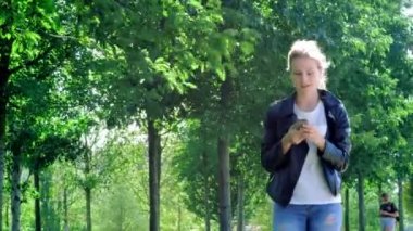 Genç güzel kız bir akıllı telefon ile parkta yürüyor. Mesajı okur ve çok mutlu. Dans ve mutluluk ve sevinç ile sıçrayan.