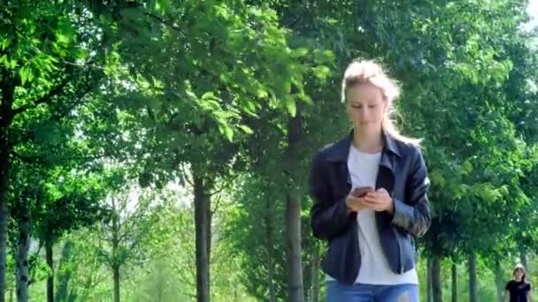 Jovem menina bonita está andando no parque com um smartphone. Lê a mensagem e está muito feliz. Dançando e saltando com felicidade e alegria . — Vídeo de Stock