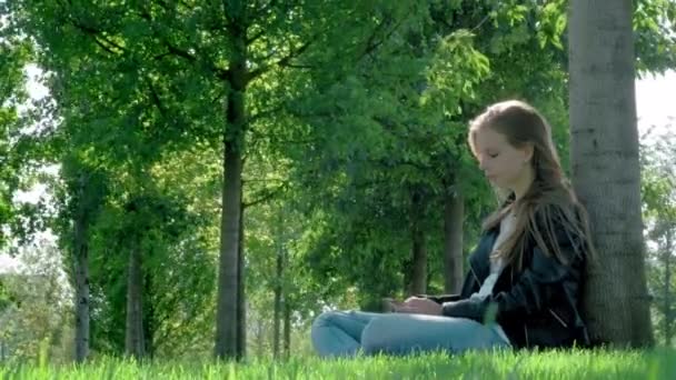 Jong mooi meisje in straat kleren in een park. Het houden van een smartphone in zijn handen, zittend op het gras onder een boom. Leest saaie boodschap en foto's, Yawns zwaar. Niet genoeg slaap, wil slapen — Stockvideo