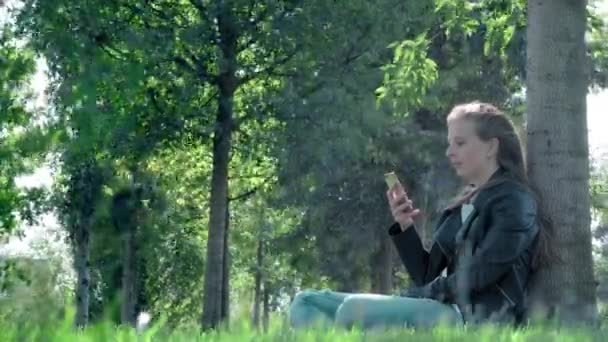 Giovane bella ragazza in abiti da strada in un parco. Tenendo uno smartphone tra le mani, seduto sull'erba sotto un albero. Legge il messaggio noioso e le fotografie, sbadiglia pesantemente. Non abbastanza sonno, vuole dormire — Video Stock