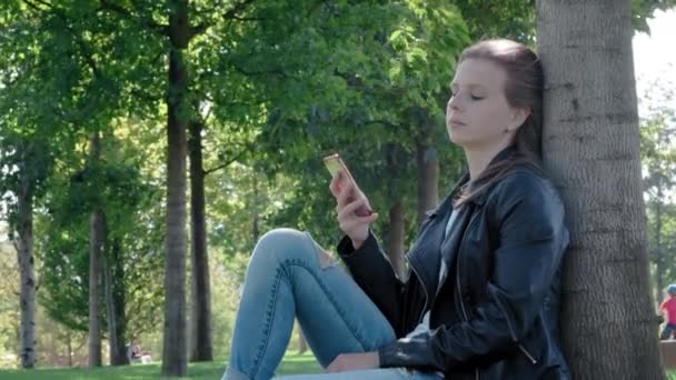 Giovane bella ragazza in abiti da strada in un parco. Tenendo uno smartphone tra le mani, seduto sull'erba sotto un albero. Legge il messaggio noioso e le fotografie, sbadiglia pesantemente. Non abbastanza sonno, vuole dormire — Video Stock