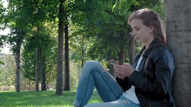 Ένα νεαρό όμορφο κορίτσι στα ρούχα του δρόμου σε ένα πάρκο. Κρατώντας ένα smartphone στα χέρια του, καθισμένος στο γρασίδι κάτω από ένα δέντρο. Γράφει και διαβάζει μηνύματα σε ένα κοινωνικό δίκτυο. Συνομιλούν με μια ομάδα φίλων — Αρχείο Βίντεο