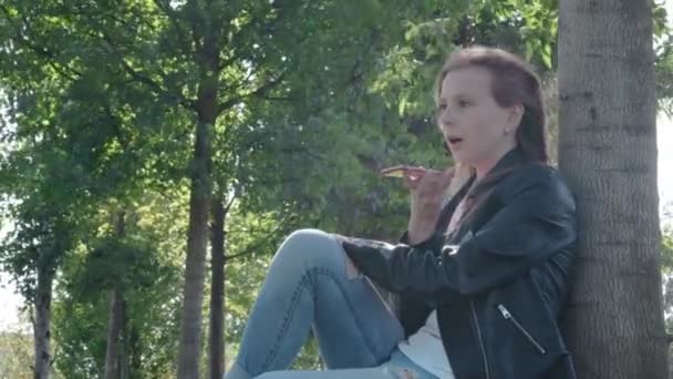 Молода красива дівчина в вуличному одязі в парку. Тримаючи в руках смартфон, сидячи на траві під деревом. Записує голосове повідомлення для друзів. Диктує текст повідомлення — стокове відео