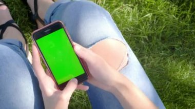 Bir parkta sokak kıyafetleri içinde genç güzel kız, yırtık kot pantolon. Elinde Yeşil ekranlı bir akıllı telefon tutuyor, bir ağacın altındaki çimlerin üzerinde oturuyor. Renk anahtarı ile Gadget, yeni teknoloji konsepti.