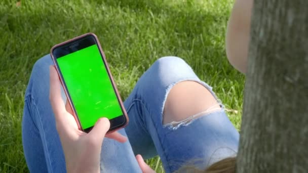 Jeune belle fille en tenue de rue dans un parc, déchiré jeans. Tenant un smartphone avec un écran vert dans ses mains, assis sur l'herbe sous un arbre. Gadget avec clé chromatique, nouveau concept technologique . — Video