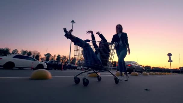 Dos novias millennials en ropa de calle se divierten en un estacionamiento de supermercados al atardecer. Montar en un carrito de compras, disfrutar de la libertad — Vídeo de stock