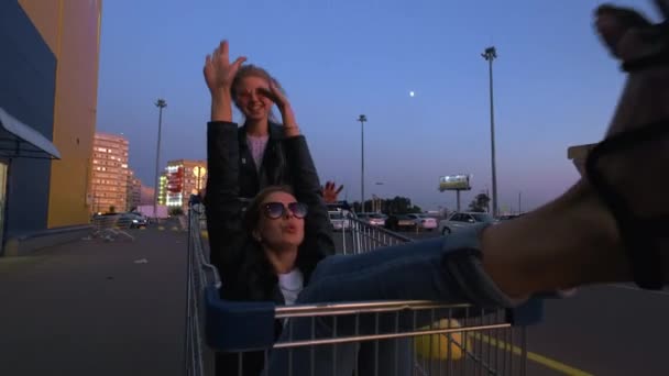 Two millennials girlfriends in street clothes have fun in a supermarket parking lot at sunset. Riding a shopping cart, enjoying freedom — Stock Video