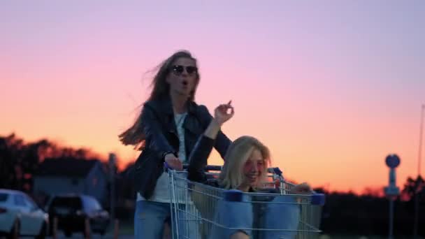 Dos novias millennials en ropa de calle se divierten en un estacionamiento de supermercados al atardecer. Montar en un carrito de compras, disfrutar de la libertad — Vídeo de stock