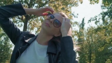 Parktaki kameraya poz veren uzun saçlı güzel genç kız modeli, yeşil ağaçların arka planında. Bir selfie için iyi bir poz arar. Deri ceket giymiş