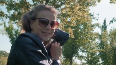 Parktaki kameraya poz veren uzun saçlı güzel genç kız modeli, yeşil ağaçların arka planında. Bir selfie için iyi bir poz arar. Deri ceket giymiş