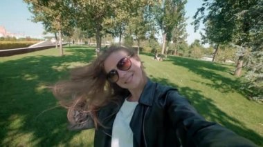 Parkta kız öğrenci. Deri ceketli sevimli kız sonbahar parkında dolaşıyor ve kendini kameraya çekiyor. Sosyal ağlar için akıllı telefonda selfie çekiyor. yavaş çekim.