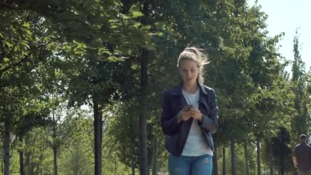 年轻的漂亮女孩带着智能手机在公园里散步。阅读消息，很高兴。快乐和快乐的舞蹈和弹跳. — 图库视频影像