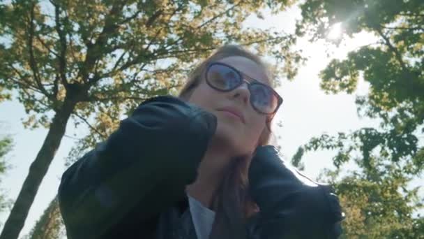 Parktaki kameraya poz veren uzun saçlı güzel genç kız modeli, yeşil ağaçların arka planında. Bir selfie için iyi bir poz arar. Deri ceket giymiş — Stok video