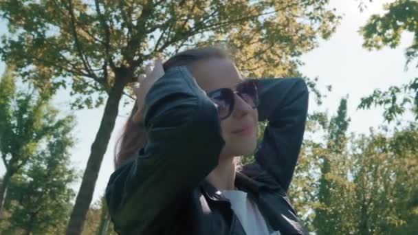 Mooi jong meisje model met lange haren poseren voor de camera in het Park, op een achtergrond van groene bomen. Zoekt naar een goede pose voor een selfie. Gekleed in een leren jasje — Stockvideo