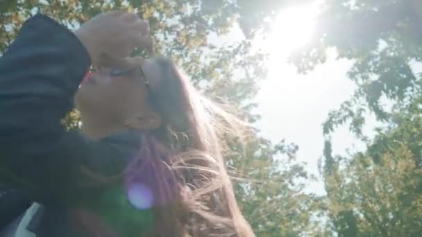 Modelo de menina bonita com cabelos longos posando para a câmera no parque, em um fundo de árvores verdes. Procura uma boa pose para uma selfie. Vestido com uma jaqueta de couro — Vídeo de Stock