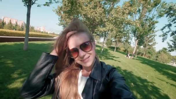 Female student in the park. Charming girl in leather jacket walks around autumn park and films herself on the camera. Makes a selfie on a smartphone for social networks. slow motion. — Stock Video