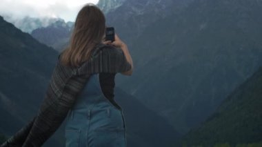 Genç çekici kız gezgin-blogger bir akıllı telefon üzerinde büyük dağların bir fotoğraf yapar. Sıcak giysiler ve ekose giymiş. Bir dağ nehri boyunca yürümek. Tetnuldi Dağı, Svaneti, Gürcistan