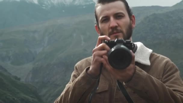 Porttait: mannelijke reiziger fotograaf maakt professionele bergen Foto's voor blog. Een man met lang haar en een baard die een professionele camera in zijn handen houdt. Expeditie naar de bergen. — Stockvideo
