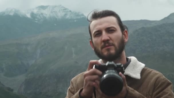 PORTTAIT: Fotógrafo viajante masculino faz fotos de montanhas profissionais para blog. Um tipo com cabelo comprido e barba a segurar uma câmara profissional nas mãos. Expedição às montanhas . — Vídeo de Stock