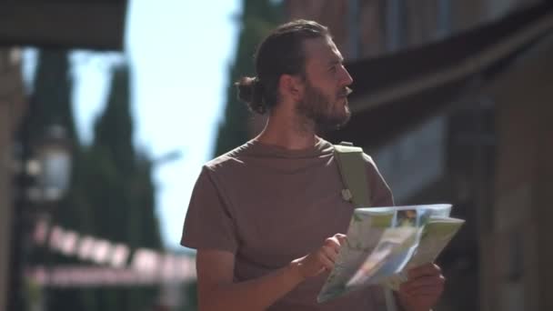 若い男の旅行者は、彼の手に地図を持ってヨーロッパの都市を歩いています。観光客は道を探しているか、失った。新しい都市の観光。地理的なアトラスを読む男の旅行者 — ストック動画