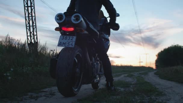 Motocyklista siedzi na motocyklu. Uruchamia rower, odjeżdża w stronę zachodzącego słońca wzdłuż wiejskiej drogi. Jedź sam. Przygody na rowerze dla kotów w przyrodzie. Romantyczny mężczyzna. — Wideo stockowe