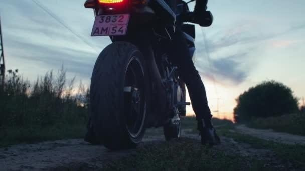 Motorrijder biker zit op een motorfiets. Start een fiets, rijdt de zonsondergang tegemoet langs een landweg. Alleen reizen. Avonturen op een kattenfiets in de natuur. Een romantische man. — Stockvideo
