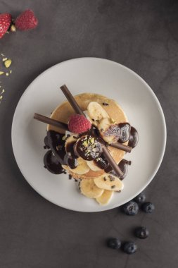 Krep çilek, ahududu, muz, çikolata, fıstık ve krem beyaz tabakta. Dikey. Boşaltmak. Üstten Görünüm