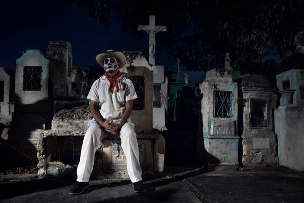 Мерида, Cementerio General, Мексика - 31 октября 2018 года: Белый мужчина в ковбойской шляпе с макияжем черепа сидит на могиле в окружении гробниц на фестивале Des Las Animas
