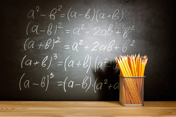 Pencils on a wooden school desk in front of a black chalkboard with Mathematical formulas school. Education concept - the desk in the auditorium