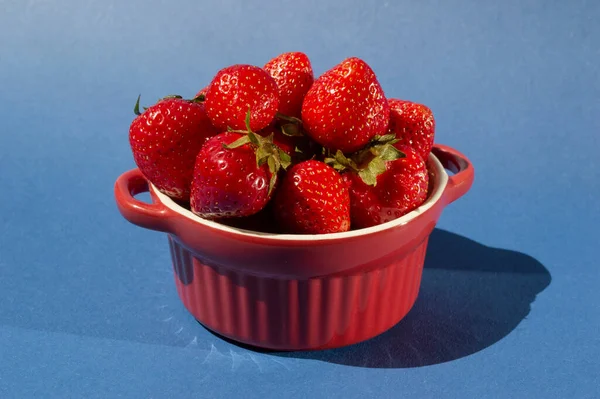 Bir grup taze lezzetli çilek kırmızı cemaric küçük tabak içinde tutacakları ile. Parlak yaratıcı çömlek konsepti. Sanat modasına uygun yaz meyveleri. Mavi arka planda güneşli gölgeler.