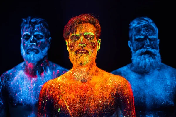 Portrait of three bearded men painted in florescent powders.