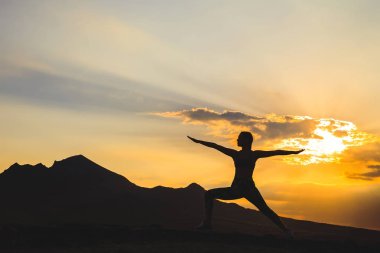 Genç kadının gün batımında veya gün doğumunda yoga ya da pilates yapan silueti, güzel bir dağ yerinde, lunge egzersizi yapıyor, Warrior ayakta.