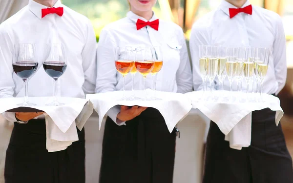 The waiters greet guests with alcoholic drinks. Champagne, red wine, white wine on trays.