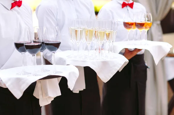 waiters greeting guests with alcoholic drinks. Champagne, red wine, white wine on trays.