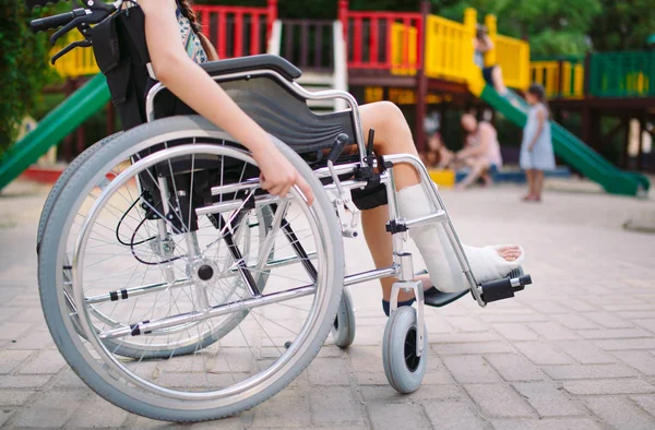 Девушка со сломанной ногой сидит в инвалидном кресле перед детской площадкой — стоковое фото