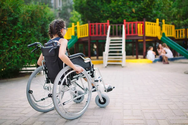 Una chica con una pierna rota se sienta en una silla de ruedas frente al patio de recreo —  Fotos de Stock