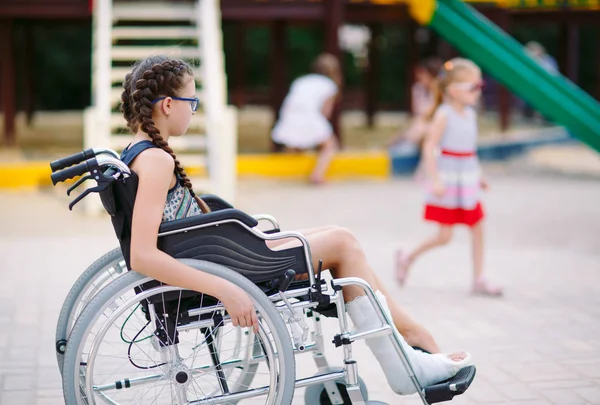 Uma menina com uma perna quebrada senta-se em uma cadeira de rodas na frente do playground — Fotografia de Stock